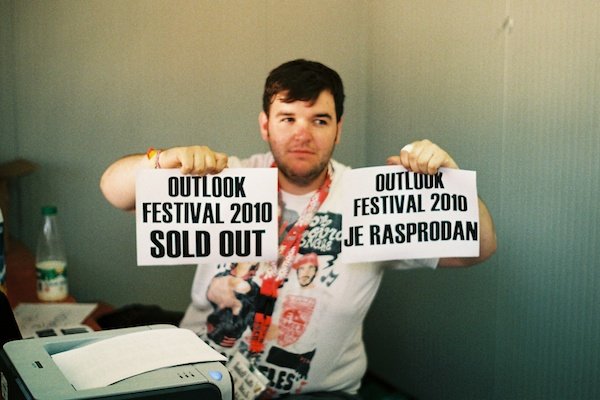Тучный мужчина держит два листа бумаги с надписью: Outlook Festival 2010 sold out