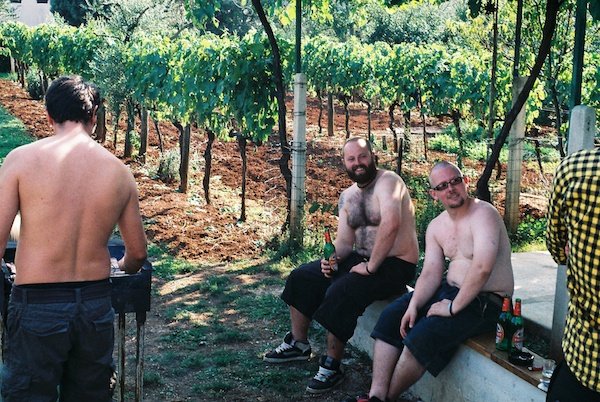 2 тучных мужчины пьют пиво рядом с виноградными лозами, а другой мужчина жарит барбекю рядом с ними
