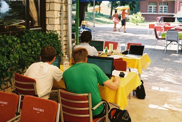 Сцена на террасе кафе, где 3 молодых человека спиной к нам работают на своих ноутбуках