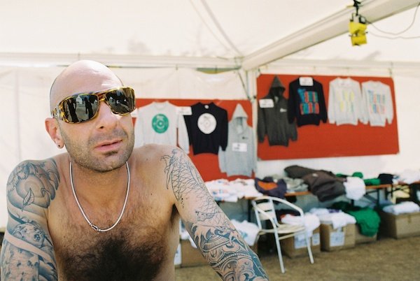 Фотография мужчины топлесс с руками, покрытыми татуировками, в солнцезащитных очках внутри торговой палатки