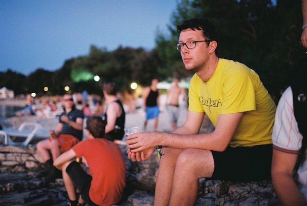 Фотография молодого человека в желтой футболке на каком-то фестивале, смотрящего в камеру