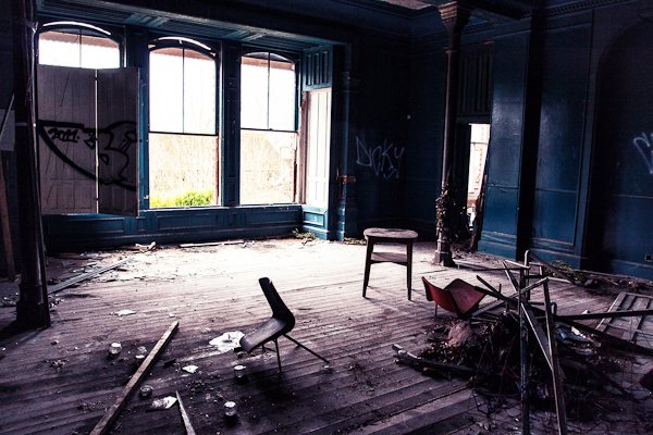 Комната в заброшенном здании с большими окнами, темными окрашенными стенами и сломанным деревом и стульями