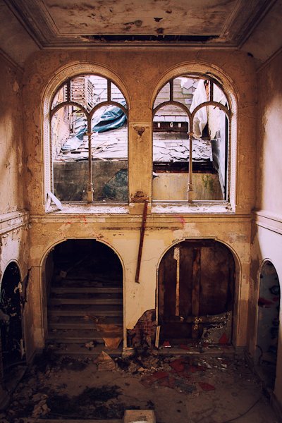 Заброшенное здание изнутри, два окна смотрят на разрушенную крышу и темную лестницу, ведущую наверх.