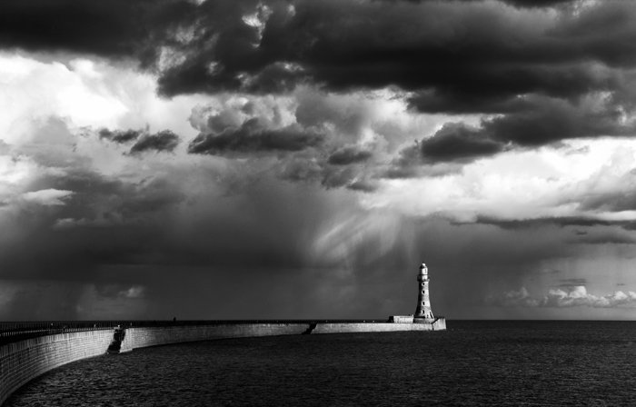 Потрясающий черно-белый снимок поцелованного солнцем маяка на мысе с темными облаками над головой