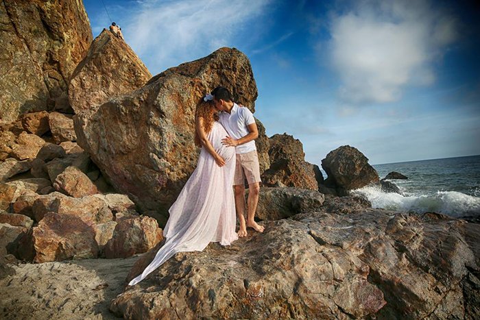 Фотография беременной пары, стоящей на скалах у пляжа. На женщине надето длинное розовое платье, и они обнимаются. 