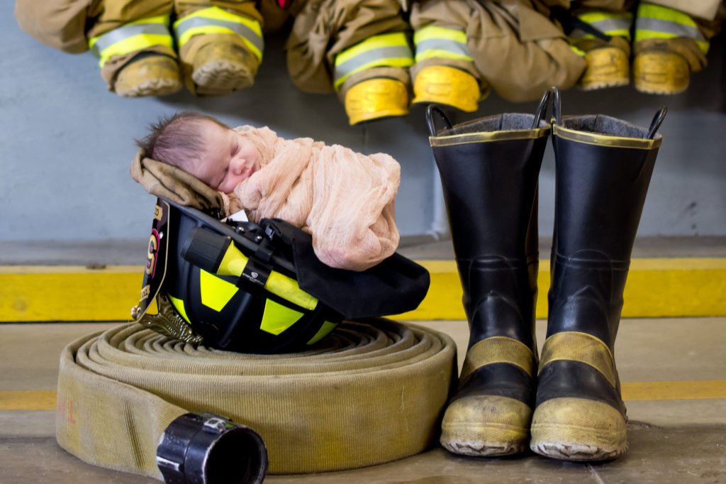 новорожденный ребенок в розовом одеяльце сфотографирован спящим в каске пожарного, в пожарном рукаве и сапогах