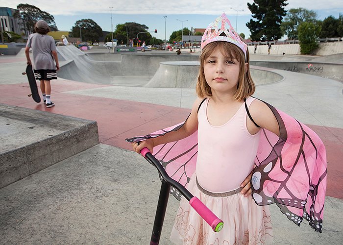 маленькая девочка в розовом костюме принцессы-бабочки со скутером, скейт-парк на заднем плане