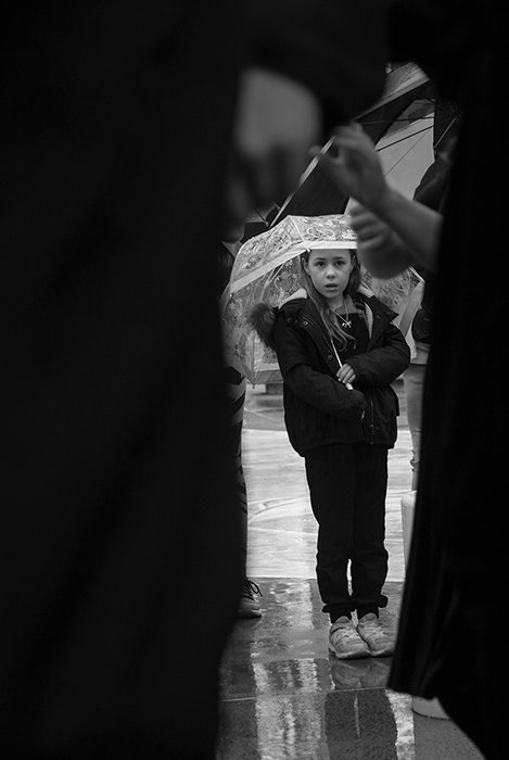Черно-белая документальная фотография маленькой девочки, держащей зонтик в обрамлении более крупных фигур