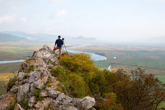 фотограф стоит на вершине скалистого холма с видом на зеленый пейзаж и реку