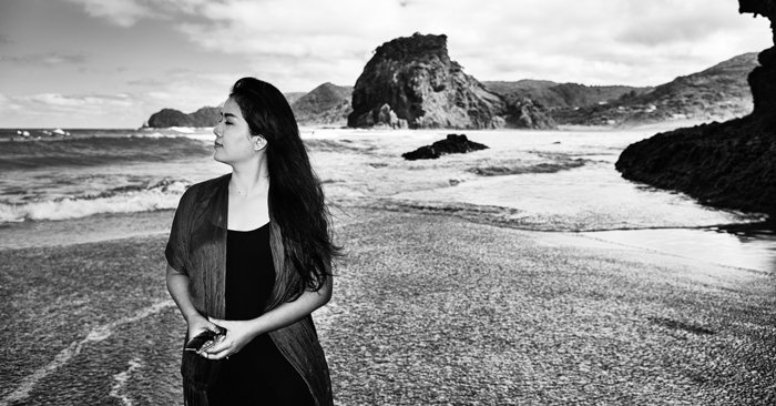 Черно-белая фотография девушки, гуляющей по пляжу Пина, Новая Зеландия, бурные волны и скалистое побережье позади нее