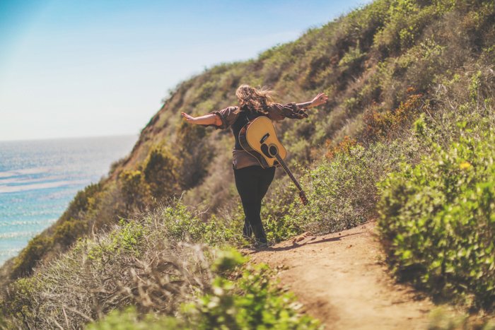 Фотография на природе человека с акустической гитарой на спине, идущего от камеры по прибрежной тропинке, вытянув руки. Улучшите свои навыки фотографии сегодня.