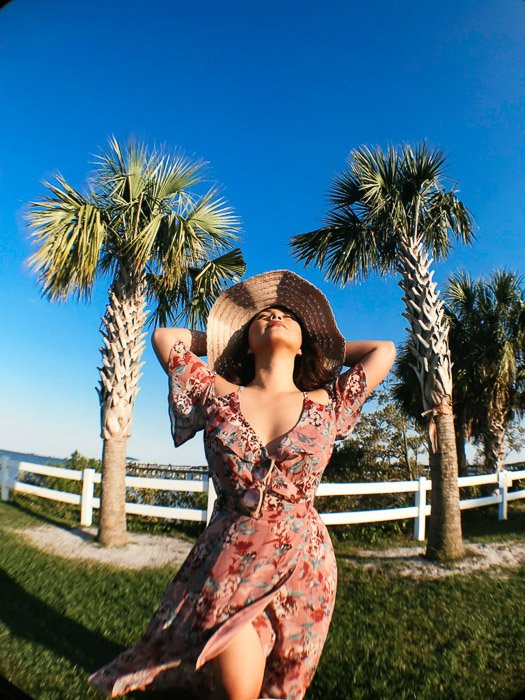 Девушка в розовом цветочном платье и соломенной шляпе стоит на траве перед белым забором и пальмами, держа руки за головой в яркий день - модная фотосессия на смартфон