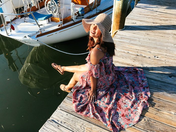 Девушка в цветочном платье и соломенной шляпе сидит на причале рядом с лодкой и улыбается в камеру. Местоположение важно для модной фотографии на смартфон