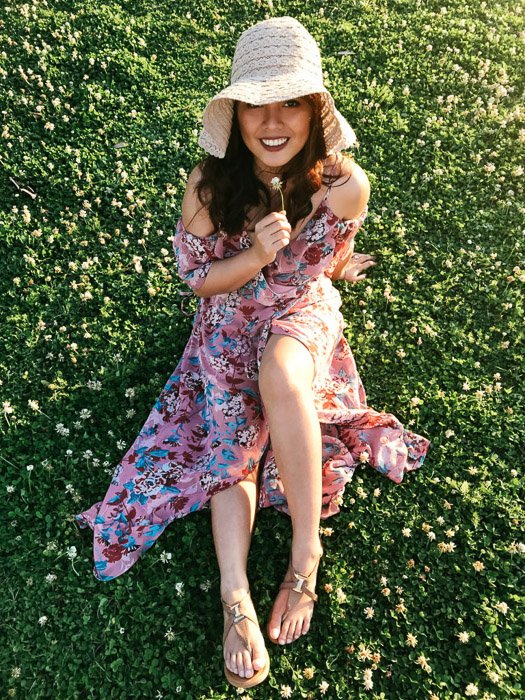 Девушка в розовом цветочном платье и соломенной шляпе сидит на траве и держит белый цветок под подбородком в яркий день - модная фотосессия на смартфон