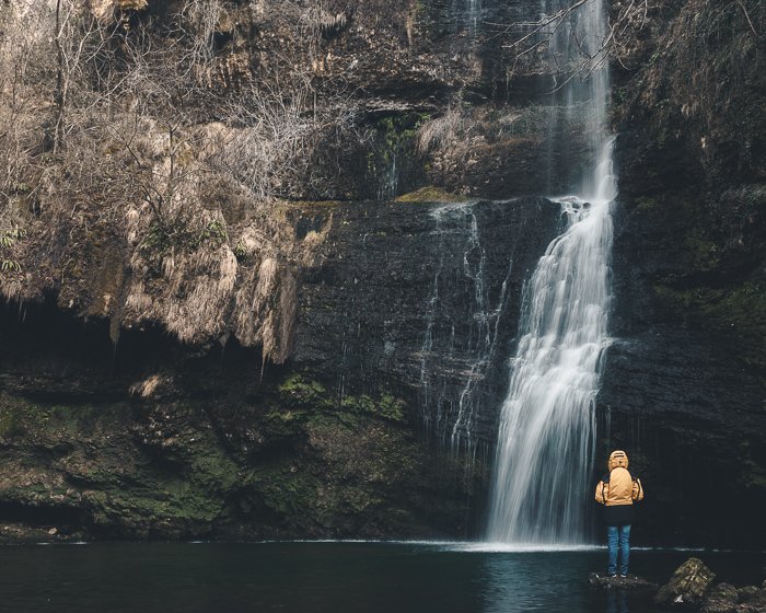 Фотография человека в желтом, стоящего перед водопадом.