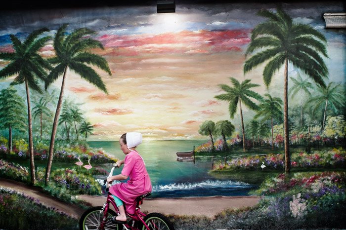Фотография девушки, проезжающей на велосипеде мимо нарисованного пейзажа от Дины Литовски. Знаменитые фотографы. 
