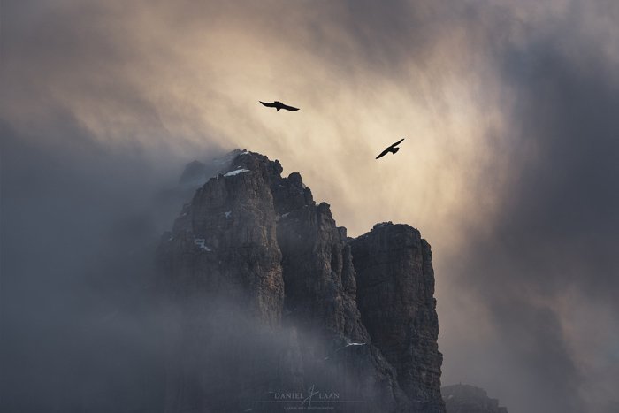 Жуткая и драматичная пейзажная фотография скалистой горы, над которой кружат птицы, и грозовое пасмурное небо.
