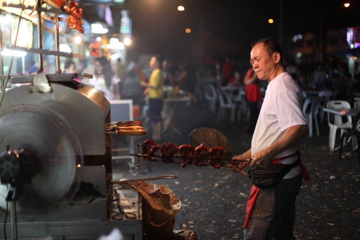 Фотография мужчины, готовящего мясо на открытом рынке при слабом освещении.