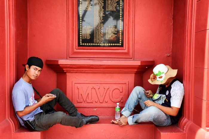 Уличная фотография двух людей, сидящих на красной стене.