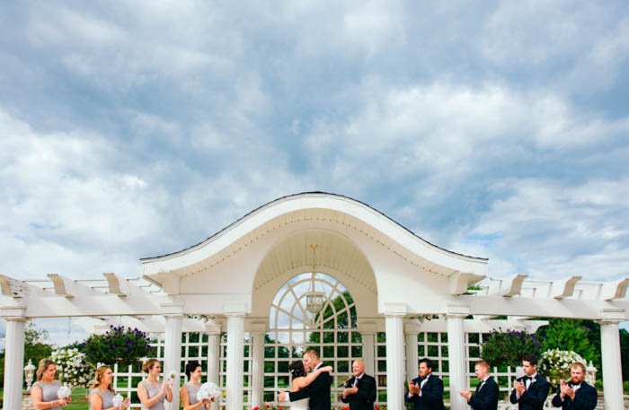 Фотография свадебной вечеринки на природе, на которой подружки невесты и жених аплодируют во время поцелуя невесты и жениха. 