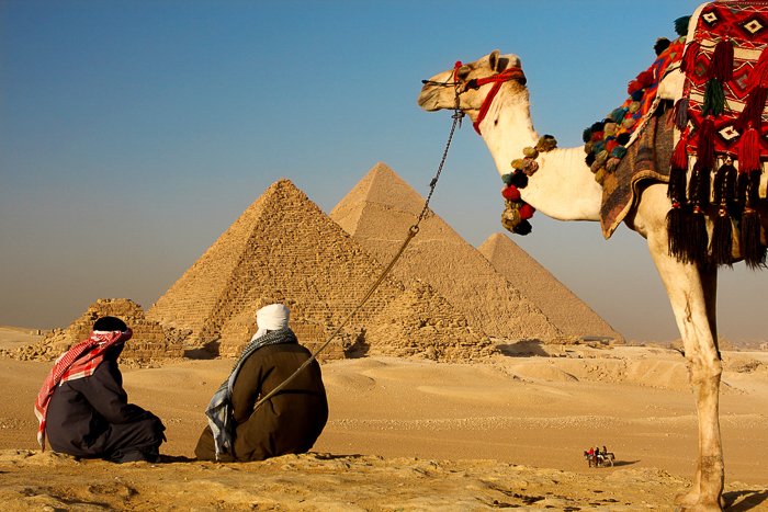 Два бедуина отдыхают на песке пустыни, глядя на пирамиды в Египте, пока их верблюд ждет