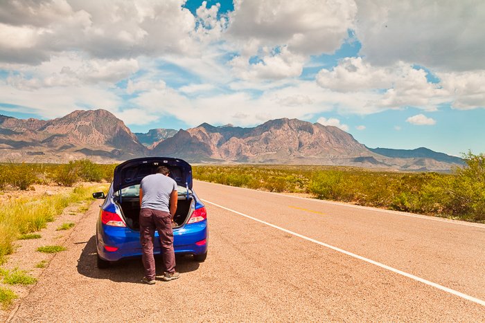 Мужчина смотрит в багажник своего синего автомобиля, остановившегося на обочине бледно-коричневой дороги, ведущей к горам вдали