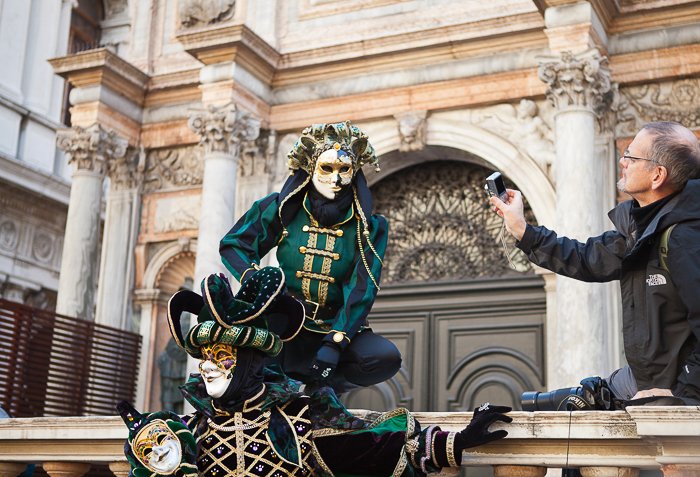 Мужчина фотографирует двух актеров в масках и костюмах, выступающих перед старым, историческим строением