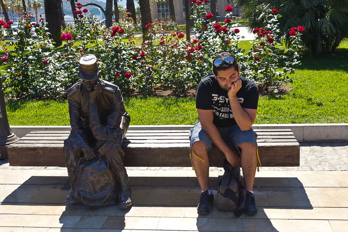 На открытом воздухе, перед россыпью красных цветов, мужчина сидит рядом со статуей, имитируя ее позу размышления