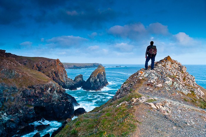 Вид сзади на человека, стоящего на вершине скалы с видом на скалистый берег и ярко-синее море и небо