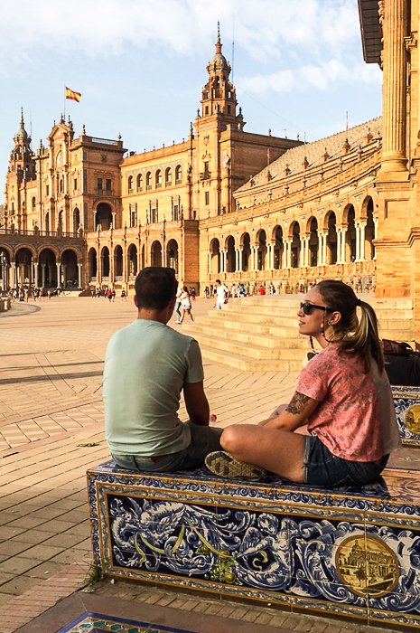 путешествующая пара сидит на украшенных ступенях в тени здания, глядя через площадь на историческую архитектуру 