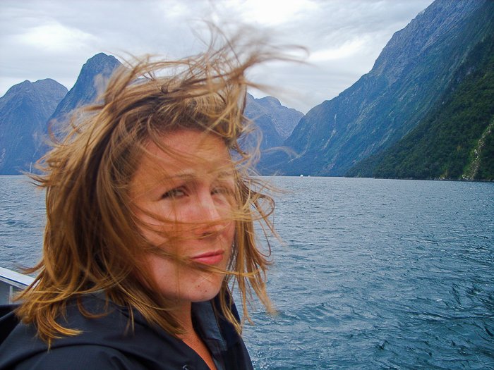 Женщина делает селфи на фоне голубого озера, неба и гор, ветер развевает ее короткие светлые волосы у лица