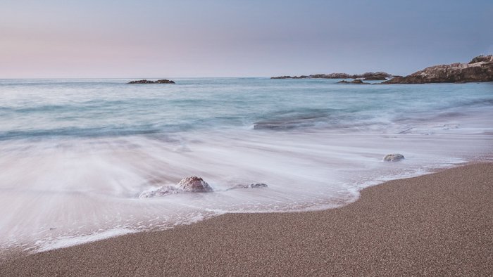 Фотография мягких пенистых волн на розоватом песке кристально синего моря на фоне пастельного восхода солнца