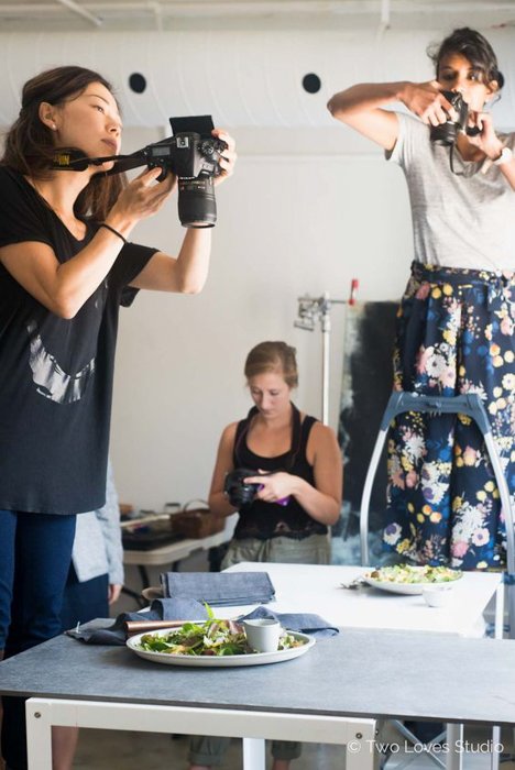 женщины с зеркальными камерами стоят на стульях, фотографируя еду на столе