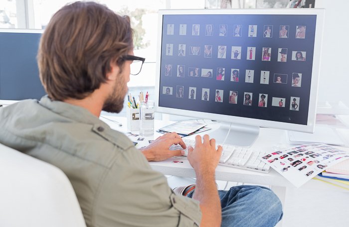 Вид сзади мужчины, сидящего за рабочим столом и просматривающего несколько фотографий на большом экране компьютера
