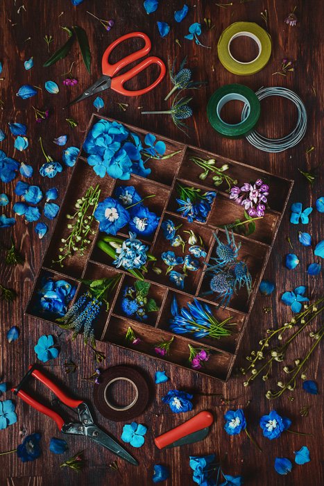 реквизит для креативных альтернативных фотофонов: небесно-голубые цветы, розовые и фиолетовые лепестки и листья в деревянной коробке, больше лепестков и листьев на фоне деревянного стола, с ножницами, щипцами и лентой