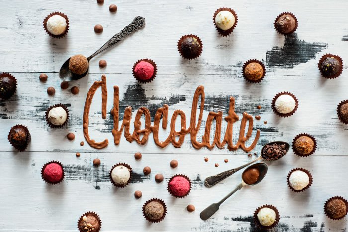 Слово шоколад из глазури с шоколадными конфетами, конфетами и чайными ложками на белом деревянном фоне. Концепция пищевой типографики. Sweet flatlay.