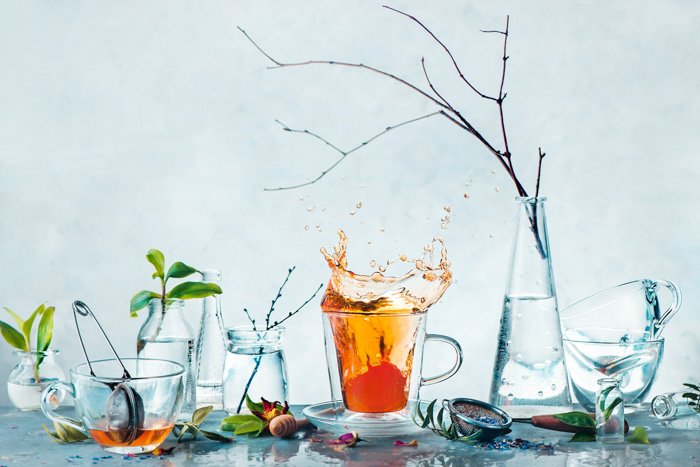Чайная чашка с динамичным всплеском и стеклянные бутылки с зелеными растениями и ветвями деревьев в весеннем натюрморте в высоком ключе. Концепция чистоты и свежести