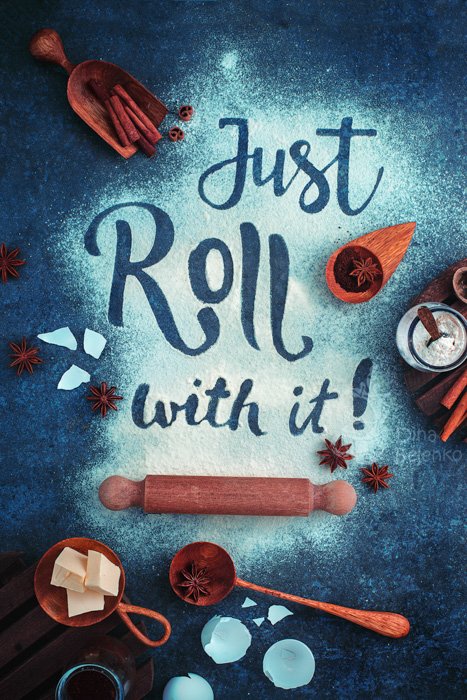 Just roll with it. Надпись в муке в окружении материалов для выпечки: скалка, деревянная ложка, ингредиенты, на темно-синем тиловом фоне