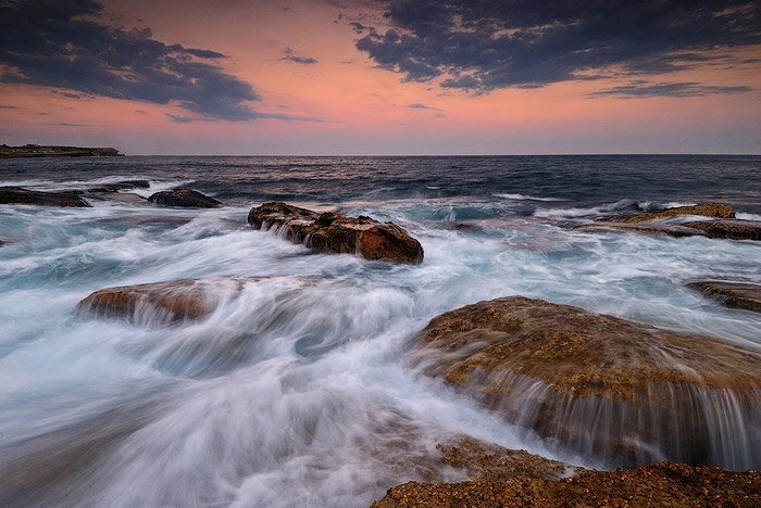 Воды, несущиеся по плоским коричневым камням на пляже Куги, Сидней, Австралия, оранжевое закатное небо на горизонте
