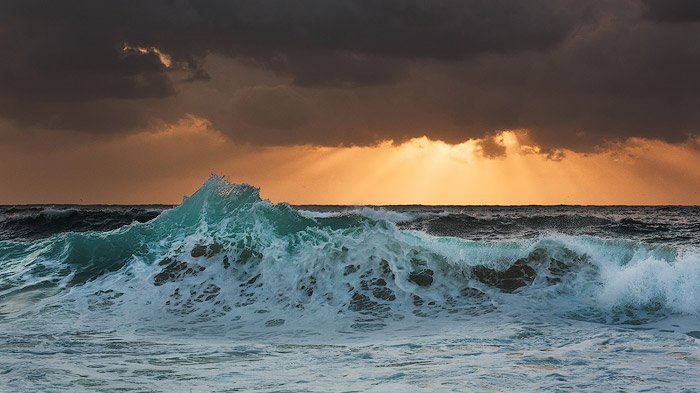 Пенистые волны разбиваются о берег, золотые лучи солнца проглядывают из-за темных облаков на горизонте. Выдержка 1/50 секунды