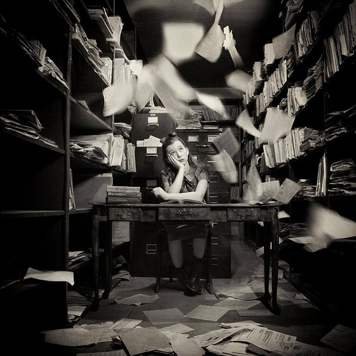 Концептуальный черно-белый портрет молодой девушки, сидящей в офисе с разлетающимися бумагами, выполненный фотографом Кэролин Хэмптон