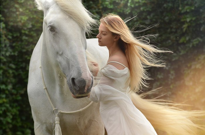 Мечтательная фотография женщины-модели с длинными светлыми волосами, позирующей рядом с белой лошадью после наложения фотошопа