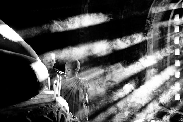 Атмосферный монохромный снимок молодых монахов, зажигающих свечи при падающем свете, проникающем через дверной проем