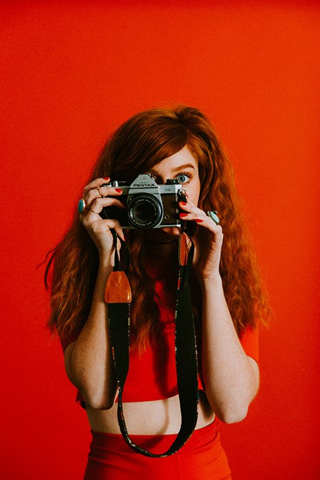 Рыжеволосая девушка перед ярко-красным экраном с фотоаппаратом Pentax