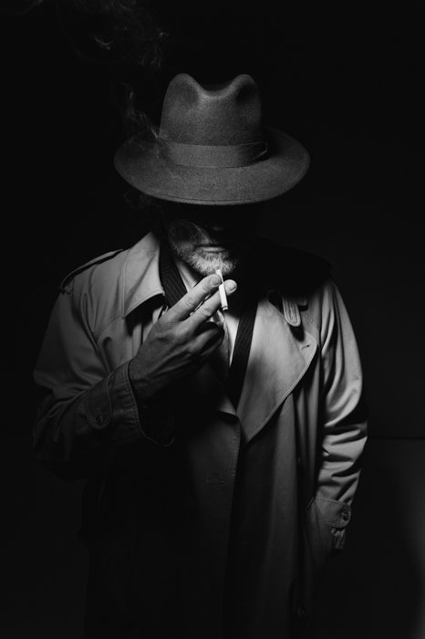 Мужчина в фетровой шляпе и плаще курит сигарету в темноте, персонаж фильма-нуар 1950-х годов