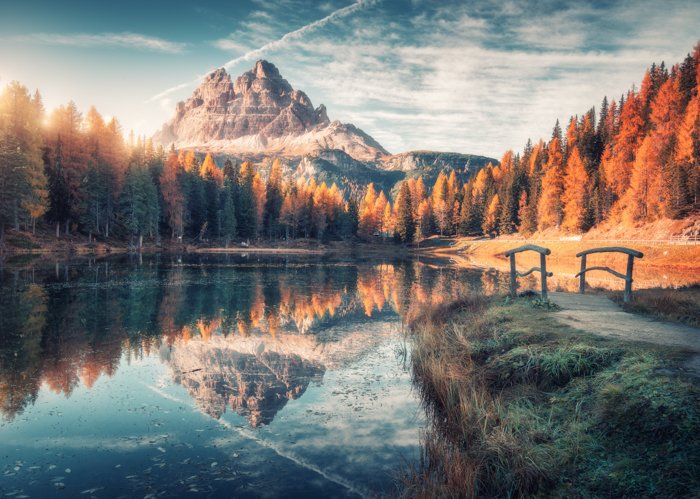 Озеро с отражением в горах на восходе солнца осенью в Доломитовых Альпах, Италия. Пейзаж с озером Анторно, небольшим деревянным мостом, деревьями с оранжевыми листьями, высокими скалами, голубым небом осенью. Красочный лес