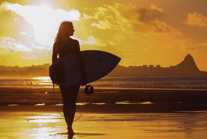 силуэт женщины, держащей доску для серфинга на пляже с подсветкой заходящего солнца