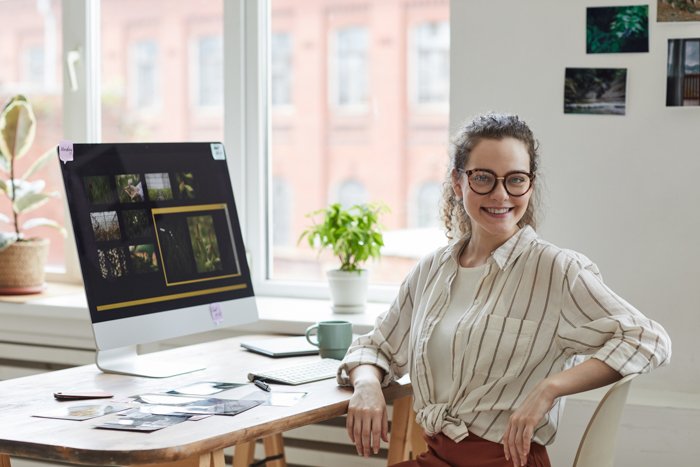 Портрет молодой женщины-фотографа, улыбающейся в камеру, позируя за столом с программой для редактирования фотографий на экране компьютера