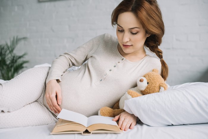 беременная женщина позирует на боку, читая книгу и держа плюшевого мишку
