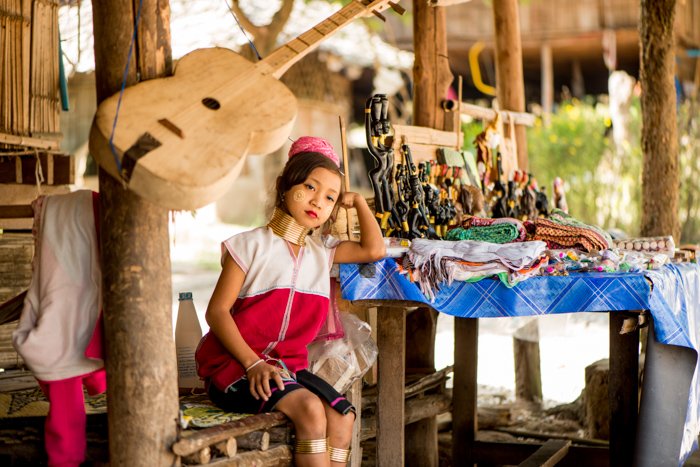 Экологическая портретная фотография молодой девушки из племени кайян, сидящей возле стола с безделушками, которые она продает возле дома семьи